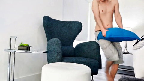 Shirtless muscular young man in gray shorts holding blue pillow in modern bedroom