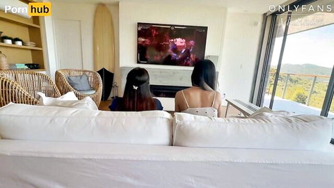 Two sexy Asian babes in skimpy tops sitting on couch watching porn TV in beach house