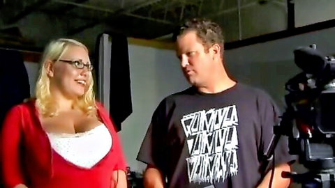 Busty blonde with glasses smiles showing deep cleavage in red cardigan on adult video set