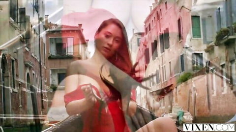 Sensual Asian redhead in revealing red dress teasing cleavage in blurred Venice streets