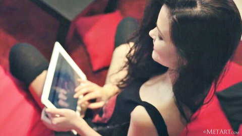 Sensual busty brunette babe showing deep cleavage using tablet on red cushions