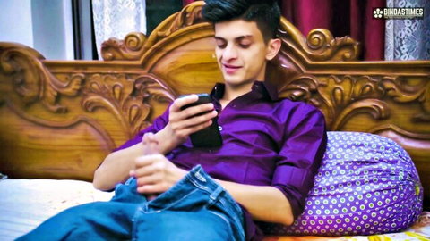Smiling Indian guy with hand down jeans stroking cock while watching phone on ornate bed