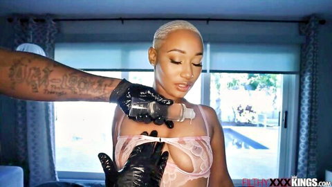 Gloved black hand pours oil on bald ebony girl's big exposed tits in pink sheer lingerie