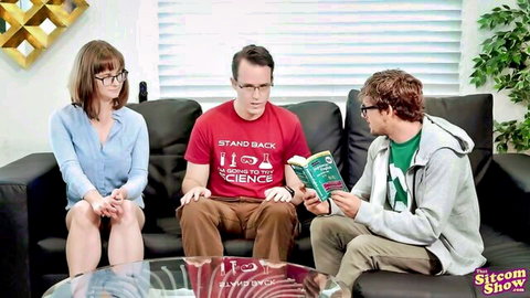 Brunette woman with glasses sits clothed between two men on couch in sitcom scene