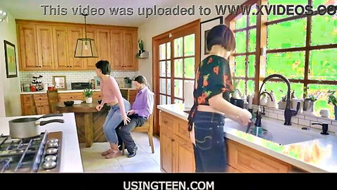 Brunette stepmom in green top washes dishes while teen in pink top teases man in kitchen taboo scene