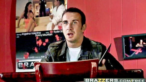 Shocked man in leather jacket sits in red chair with Brazzers posters of Angela Valentina and Rudy Mountain behind him