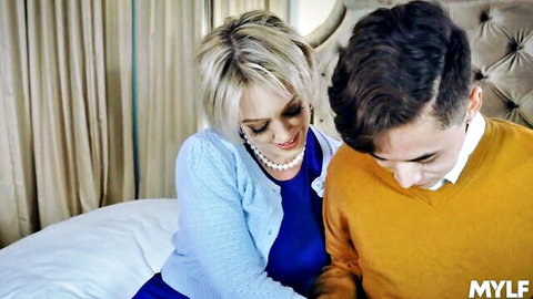Smiling blonde MILF in blue dress leans close to young stud in sweater on bed