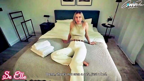 Blonde babe in cream halter top and wide pants sits legs spread on hotel bed teasing cleavage