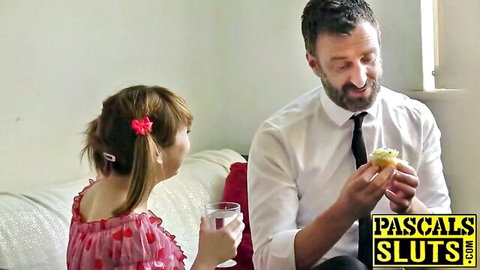 Pigtail teen girl in pink dress holds water glass as suited man offers cupcake smiling on couch