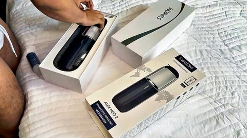 Man's hairy hand unboxing SVAKOM Alex Neo 2 black male masturbator stroker toy from white box on bed