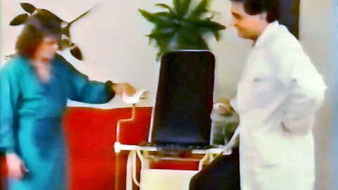Vintage doctor in white coat stands with woman in blue robe by gyno chair holding white cloth