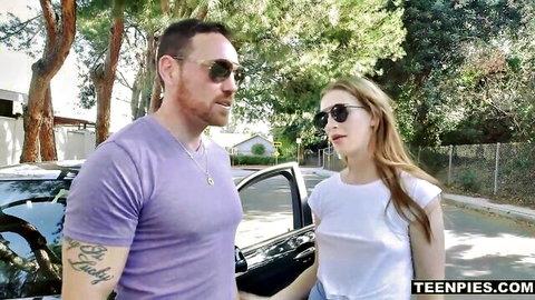 Blonde teen in white top smiles with tattooed guy by black car outdoors