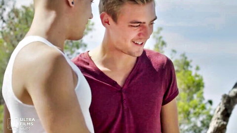 Two young fair-skinned men smiling closely outdoors, one in white tank top and one in maroon shirt