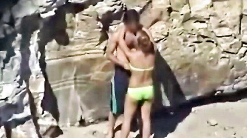 Shirtless guy passionately hugs redhead babe in neon green bikini from behind on rocky beach
