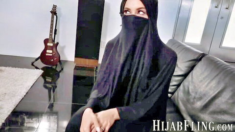Seductive niqab woman in black hijab smokes teasingly with piercing eyes on leather couch