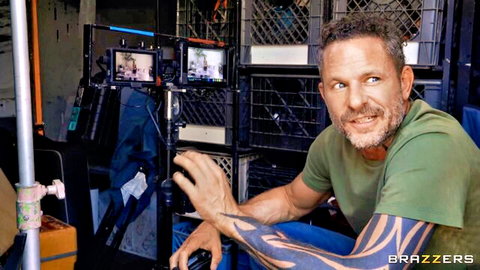 Tattooed muscular man in green shirt smiles on Brazzers porn set with studio monitors and crates