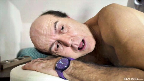 Bald old man lying naked on bed with purple watch mouth open in ecstasy