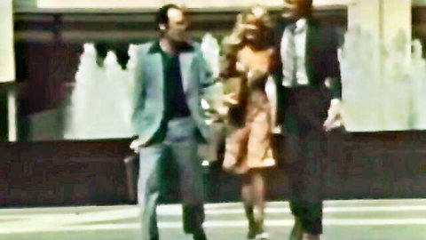 Vintage 1976 porn thumbnail: blonde in orange dress walks with suited men by fountain