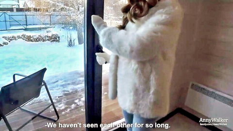 Blonde in fluffy white fur coat poses seductively by snowy window overlooking winter landscape