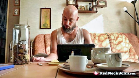 Bald bearded man in tank top sitting at table using laptop with tea cups and coin jar in cozy room
