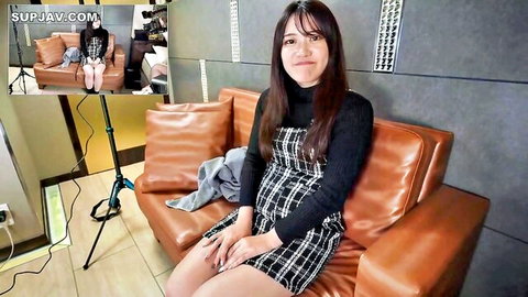 Cute Japanese college girl in black turtleneck and plaid skirt sits smiling on leather couch