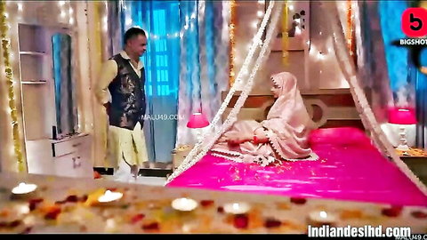 Hot Indian bride in white saree lying seductively on pink wedding bed with husband in kurta standing nearby