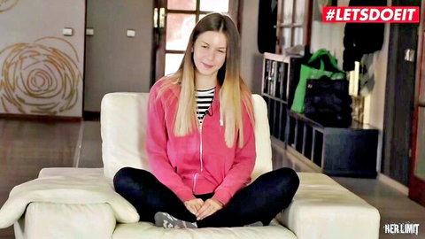 Young blonde babe in pink hoodie and black leggings sits smiling on white couch
