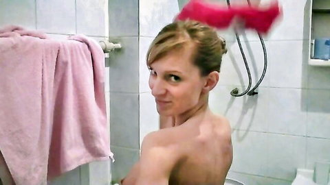 Playful blonde woman nude in bathroom, smiling over shoulder while peeing