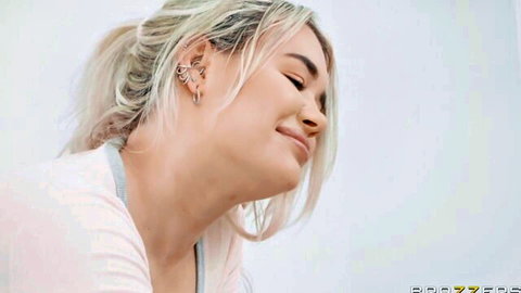 Gina Valentina smiles teasingly in pink top, blonde ponytail and earrings