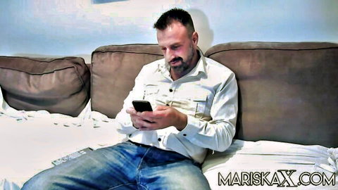 Bearded man in white shirt and jeans sits on couch checking phone casually