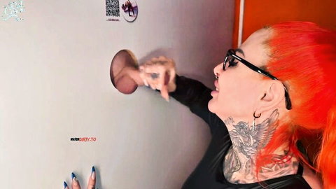 Tattooed redhead with glasses sticks hand through glory hole in wall