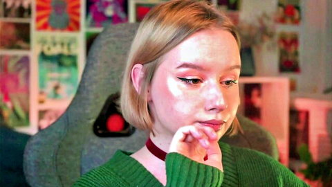 Blonde webcam girl in green sweater and choker poses thoughtfully in gaming chair