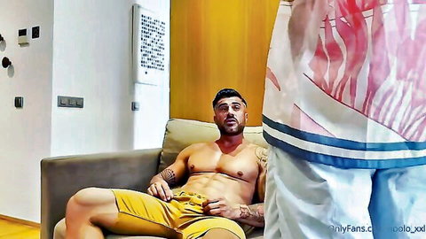 Muscular tattooed hunk sits on couch with shorts pulled down exposing thick cock base as buddy bends over in gay scene
