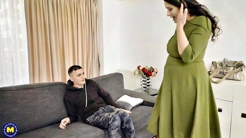 Mature woman in green dress talks on phone standing over young man lounging on couch
