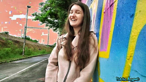Brunette beauty in trench coat smiling against graffiti wall in public street outdoor teasing