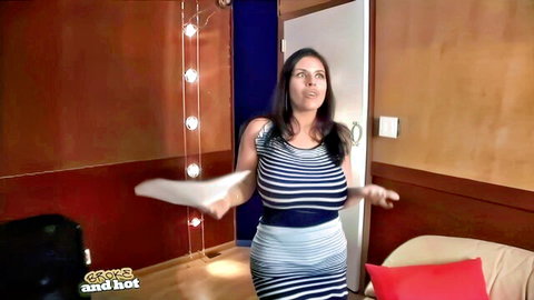 Curvy brunette MILF with huge tits in tight striped dress poses with arms outstretched teasing her big boobs