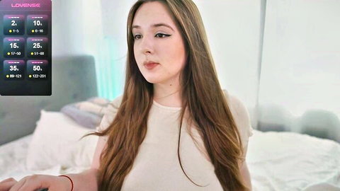Cute teen webcam girl with long brown hair smiling in loose white top on bed