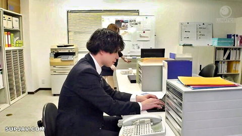 Suited young Japanese man typing at office desk while brunette colleague leans behind him