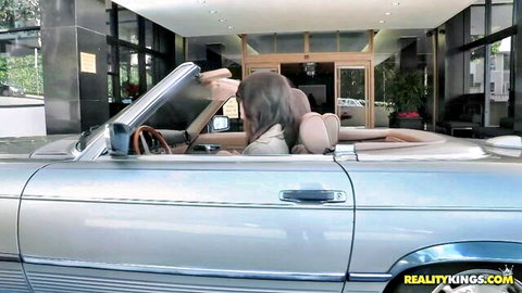 Tanned brunette babe exposing medium tits sitting topless in silver convertible outside hotel