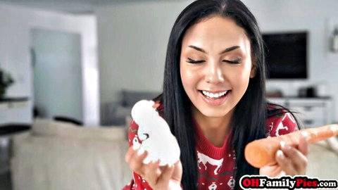 Smiling brunette babe in red Christmas sweater holding thick white and orange dildos teasingly