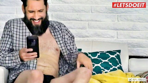 Bearded man smiling in open plaid shirt and black underwear checking phone on couch