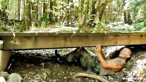 Muscular tattooed soldier in camo pants crawls under wooden bridge in forest gripping beams tightly