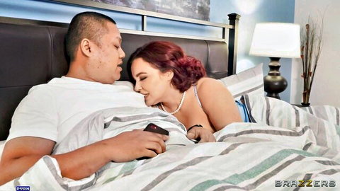 Busty redhead MILF in lingerie cuddles muscular Asian stud in bed watching phone