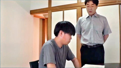 Clothed Japanese older man stands behind young guy studying at wooden table