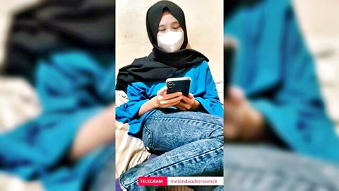 Southeast Asian hijabi teen in tight blue jeans and mask teasing on bed with phone