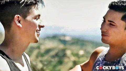 Two muscular gay studs in tank tops smiling intimately outdoors with mountain view