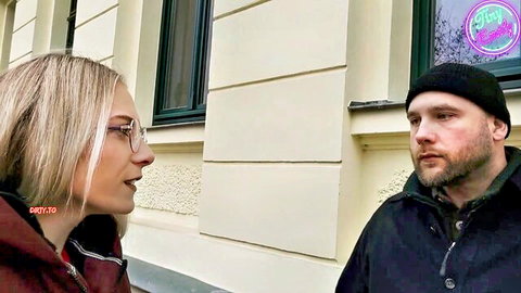 Blonde woman with glasses talks intimately face-to-face with bearded man in beanie outside building