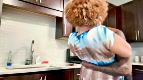 Thick ebony woman with curly hair hugged from behind by black man in kitchen