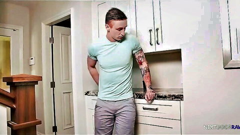 Young tattooed skinny guy in tight cyan shirt and gray pants leaning on kitchen counter teasing bulge