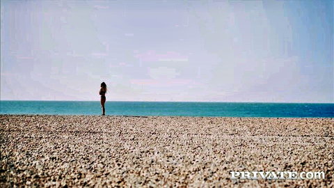 Jolee Love in black bikini on sunny pebbly beach, teasing pose